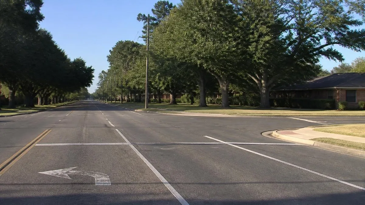 An empty tree-lined four-lane boulevard intersection with a residential side street on a sunny afternoon, representing Crestwood Boulevard in Birmingham, Jefferson County, Alabama.